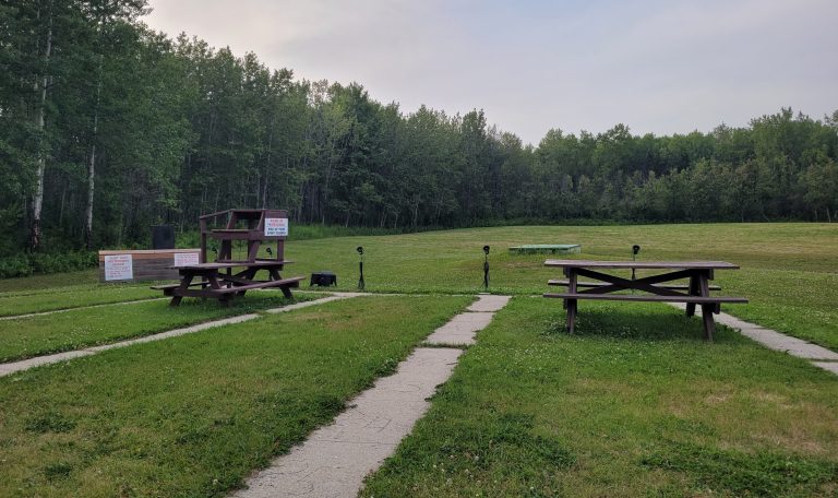 A picture of the trap shotgun area. The picture shows a wide open area with at least five spots in a curved row for the shotgun users to stand when using their weapons, with a stand for someone else to sit located in the center of the five spots and behind them. There are also two picnic benches shown sitting behind where the shotgun users would stand. To the left is a shotgun pattering board with a sign saying 1. No steel shot, 2. No shot larger than #4, and 3. No slugs. There is also another sign for the patterning board, however it is not fully shown in the image. 