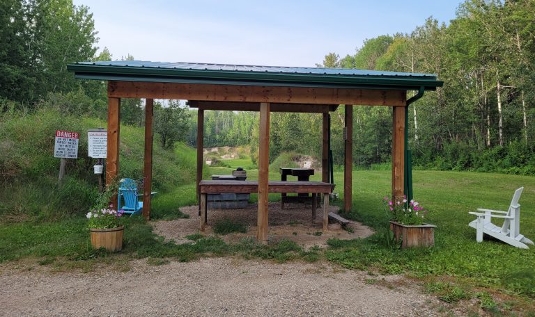 A photo of the long gun range. It shows a covered area with a table and two shooting stands. To the left and right of the covered area is a lawn chair. In the distance past the covered area three different target areas can be seen.