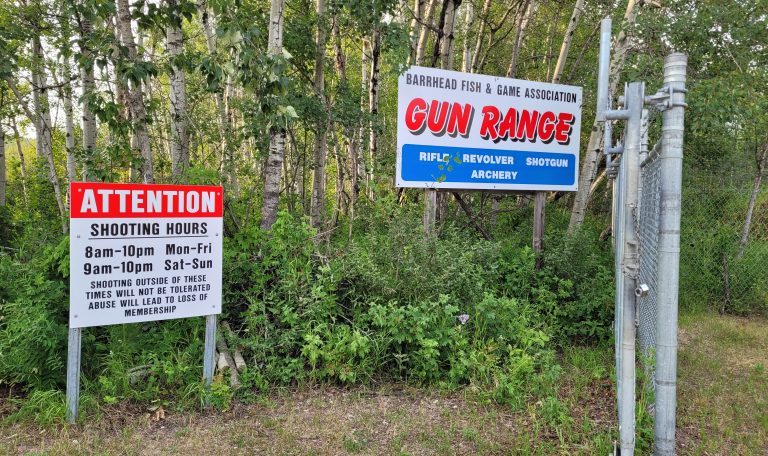 A photo showing a forested area with two signs located at the entrance to the Barrhead Fish and Game Association shooting range. One sign reads Barrhead Fish & Game Association Gun Range, and lists that it allows rifles, revolvers, shotguns, and archery. The second sign lists the open hours for the range as being 8 am to 10 pm for Monday through Friday, and 9 am to 10 pm for Saturday and Sunday. This sign also states that using the range outside of those hours will not be tolerated and will result in a loss of membership.